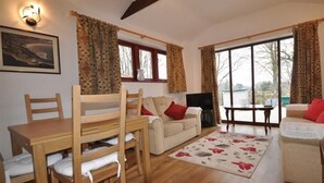 Un salon chaleureux avec une table à manger en bois, deux chaises en bois, un canapé beige agrémenté de coussins rouges, un téléviseur et une grande fenêtre avec des rideaux.