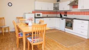 A compact kitchen with white cabinets, a microwave, an oven, and a sink. There is a dining table with chairs and a clock on the wall. - Tynk (Padstow)