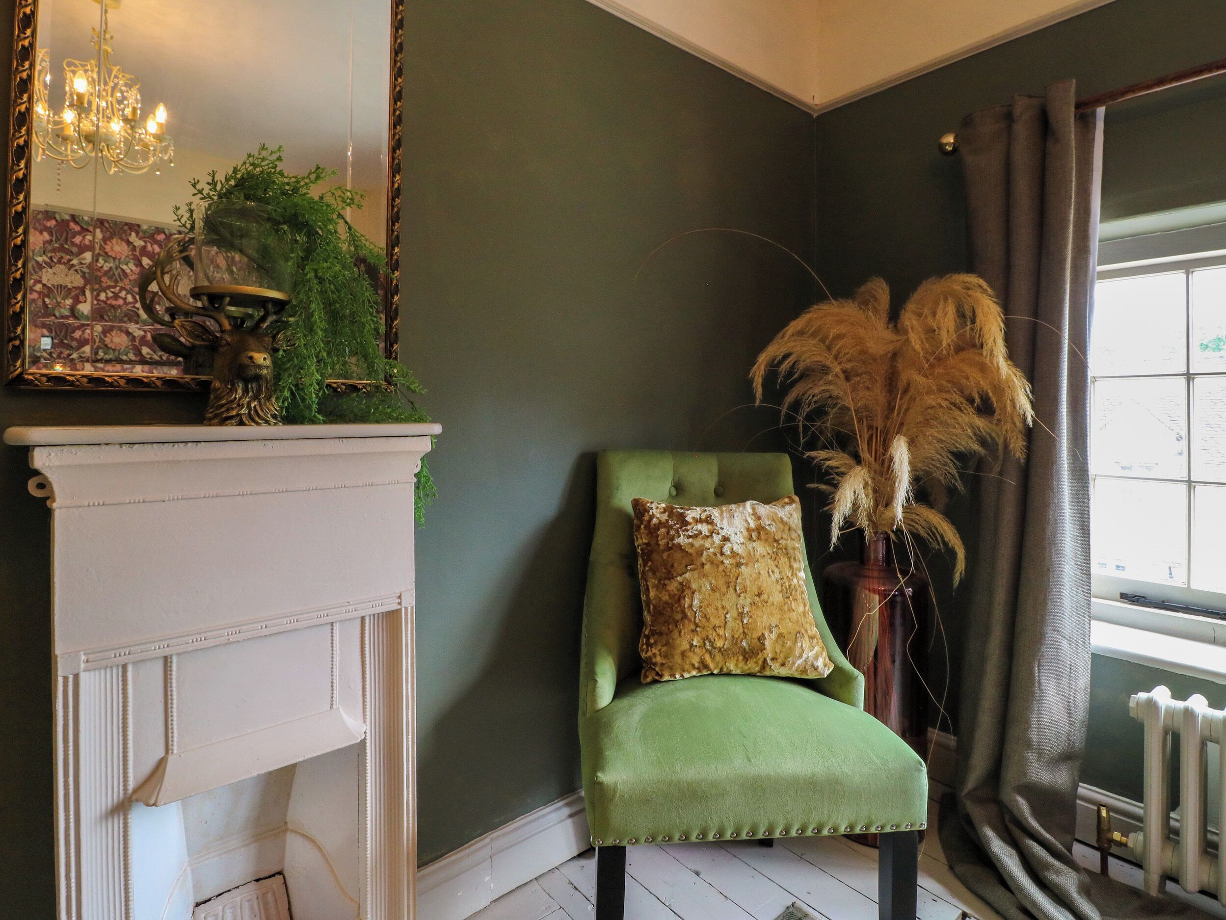 A room with a green upholstered chair, a white fireplace, and a mirror with a floral pattern.