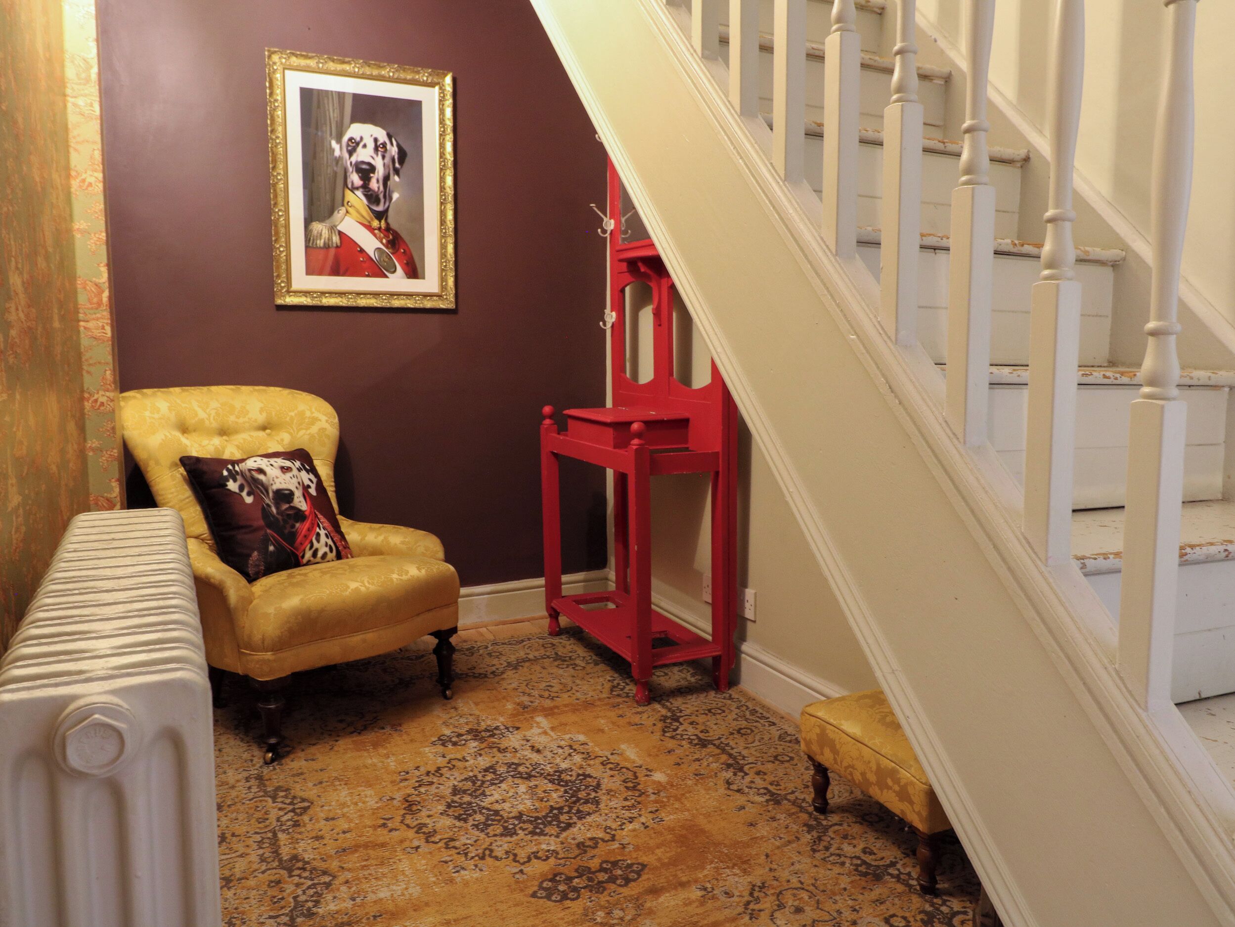 Una habitación con alfombra estampada, un sillón amarillo con un cojín de lunares de perro dálmata, una mesita auxiliar roja y un cuadro enmarcado en la pared.