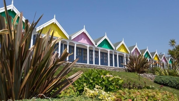 A row of colorful cottages with white trim and pitched roofs, surrounded by lush greenery. - Dream View (Weymouth)