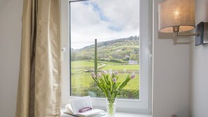 Ett sovrum med ett fönster som vetter ut mot ett grönskande landskap, ett sängbord med en bok och en vas med blommor samt en vägglampa.