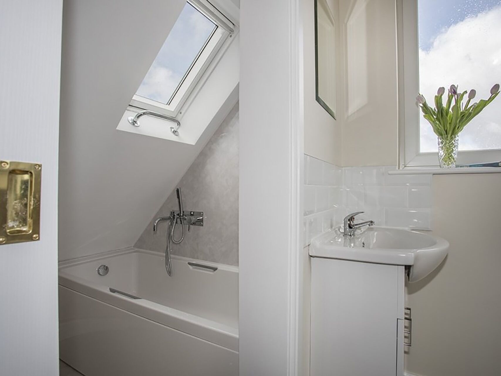 A bathroom with a skylight, a bathtub, a sink, and a window with a vase of flowers.