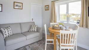 Ett mysigt vardagsrum med en grå soffa, ett matbord i trä med stolar, ett fönster med gardiner och inramade tavlor på väggen.