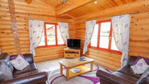 A log cabin interior with two leather sofas, a wooden coffee table, a TV on a stand, and large windows with floral curtains.