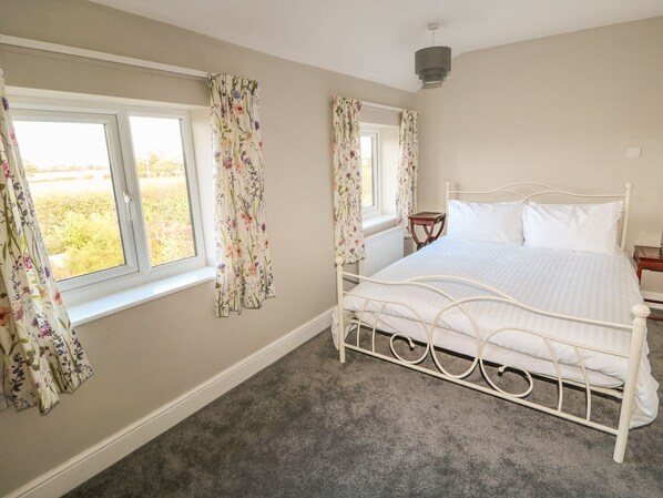 Une chambre à coucher avec un lit en métal blanc, une petite table de chevet et une fenêtre ornée de rideaux à motifs floraux.