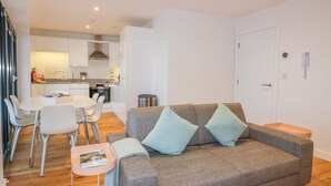 A modern living room with a grey sofa, two light blue cushions, a small table with a magazine, and a dining area with a white table and chairs. - Onshore 7 (Newquay)