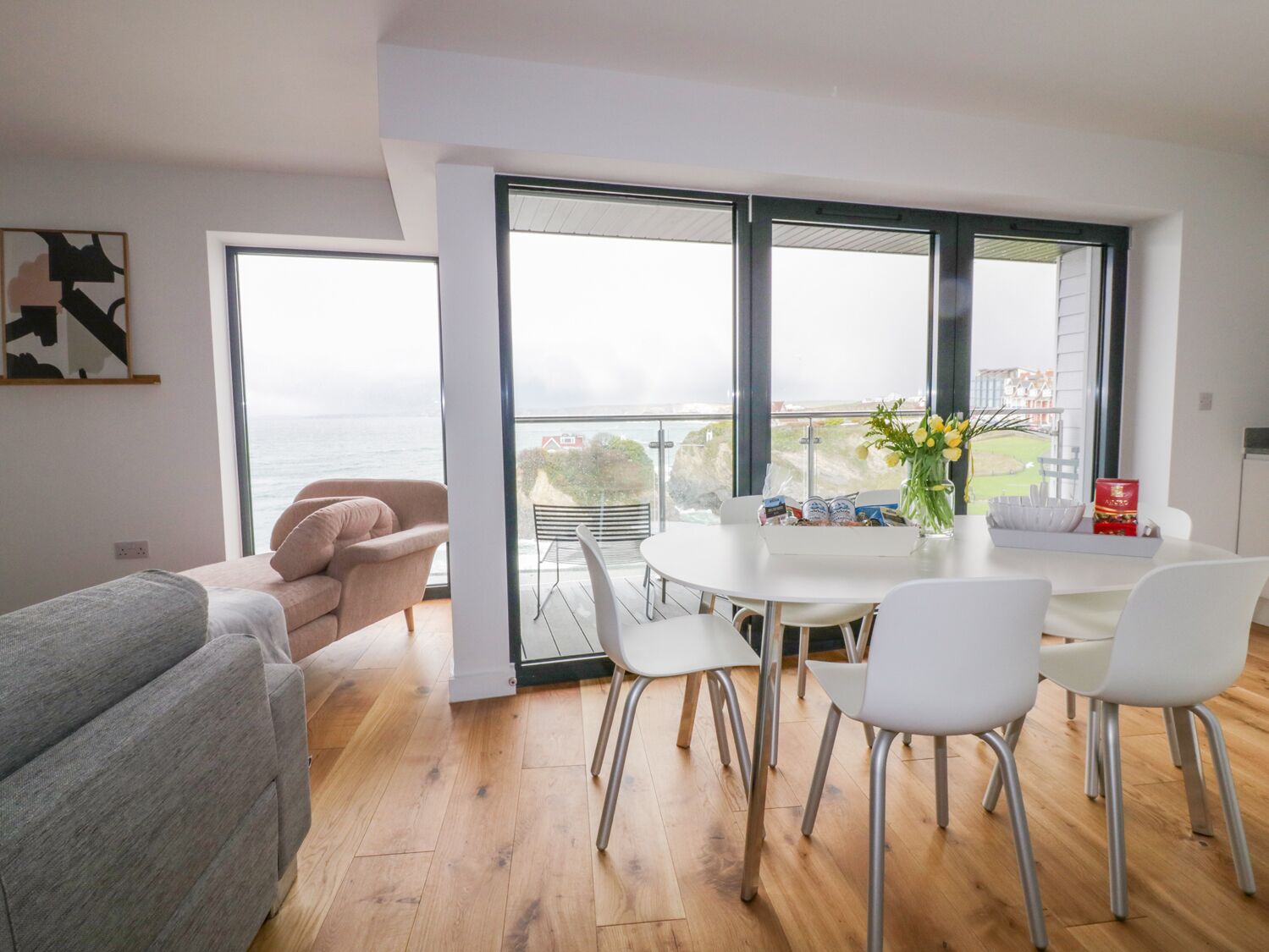 Una sala de estar moderna con un ventanal grande con vista al mar, un comedor con sillas blancas y una mesa, y un sofá gris.
