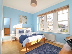 A bedroom with a bed, bedside tables, a dresser, a large window, and a view of buildings outside. - Hartland View (Woolacombe)