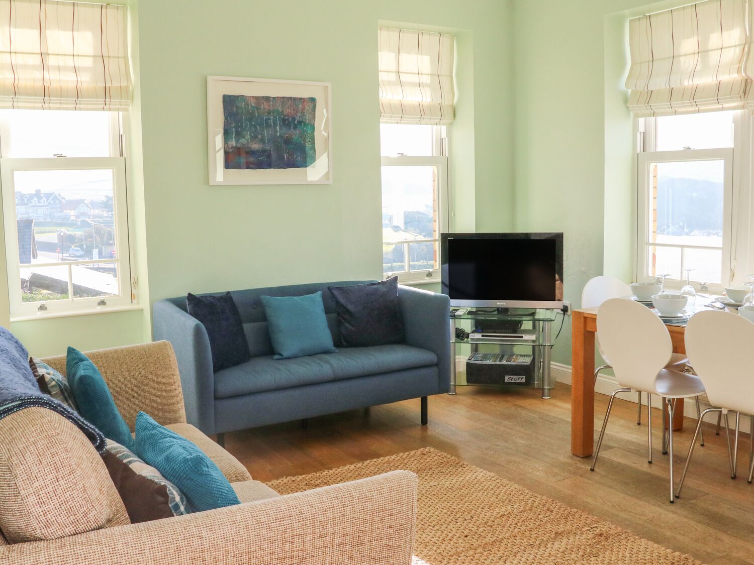 A living room with a sofa, a TV on a glass stand, a dining table with chairs, and a painting on the wall.