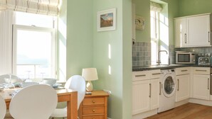 A kitchen with white cabinets, a microwave, a washing machine, and a dining area with a table and chairs. - Hartland View (Woolacombe)