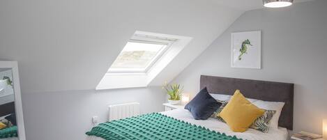 A bedroom with a skylight, a bed with a green and white bedspread, a bedside table with a lamp, and a wall-mounted plant.
