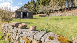 Une petite maison en pierre, avec la porte ouverte, entourée d’un mur de pierres et d’un champ herbeux.