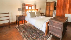 A bedroom with a wooden bed, a nightstand, a dresser, a window with curtains, and a radiator. - Cymryd Uchaf (Conwy)