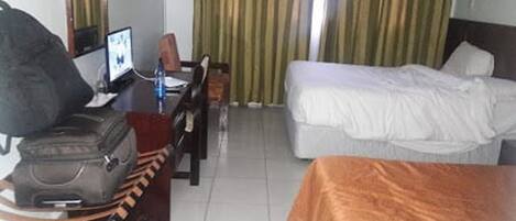 A hotel room with a bed, a desk with a computer, a chair, and a suitcase.