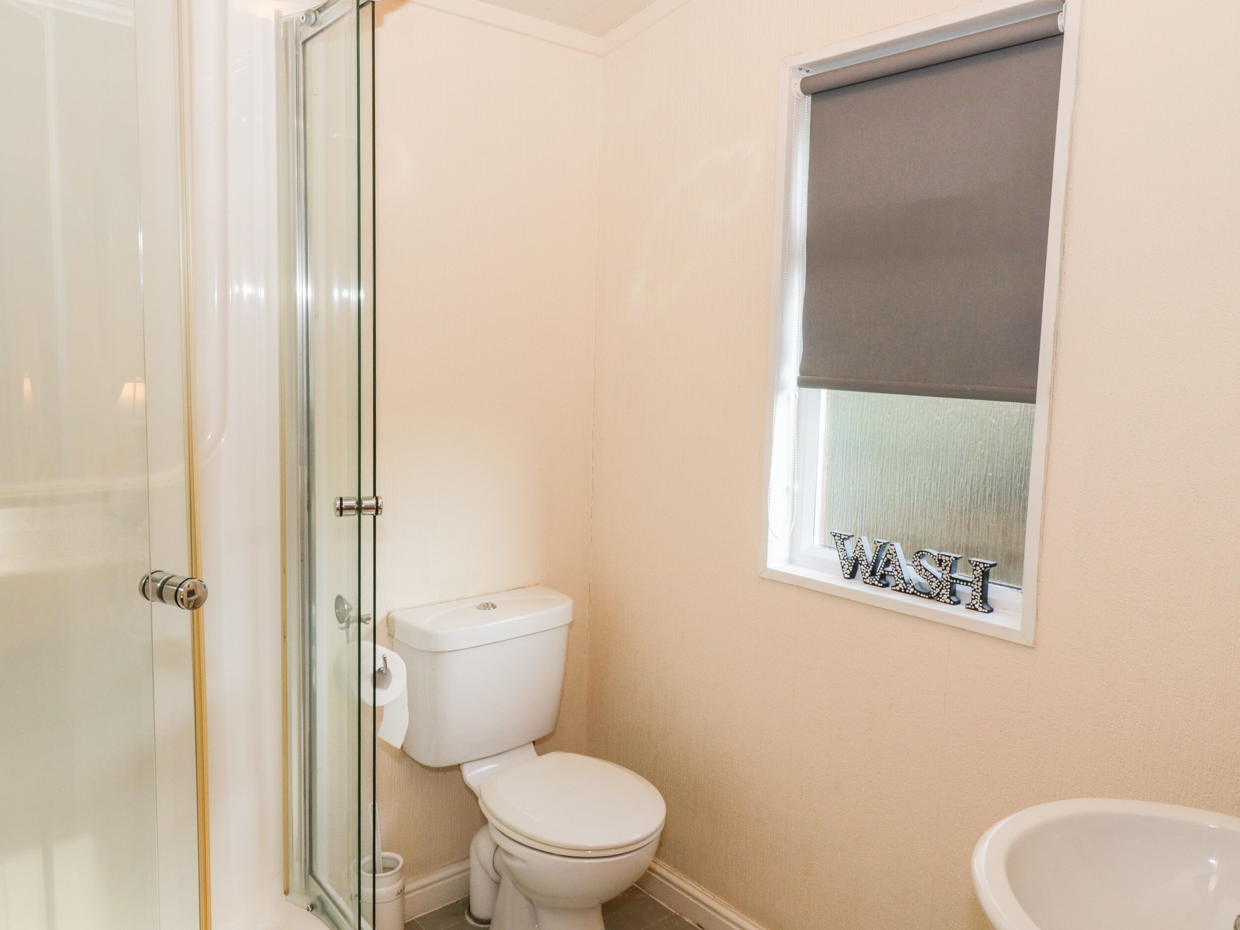 Une salle de bain comprenant des toilettes, une douche avec une porte en verre, une fenêtre sur laquelle est apposée une pancarte indiquant « LAVER », et un lavabo.