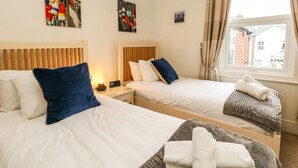 A hotel room with two beds, each with a headboard, a bedside table with a lamp, and a small picture frame. There is a window with curtains, and a view of buildings outside. - Quay Cottage (Weymouth)