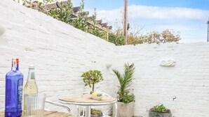 A small outdoor patio with a round wooden table and metal chairs, surrounded by white brick walls and potted plants. - Quay Cottage (Weymouth)