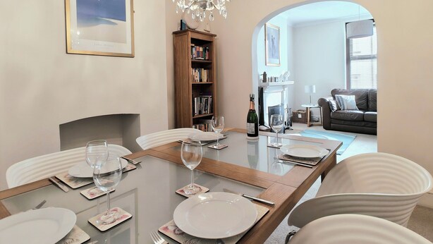A dining area with a glass table set for four, surrounded by white chairs. A chandelier hangs above, and a bookshelf is visible to the left. - Quay Cottage (Weymouth)