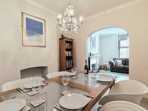 A dining area with a glass table set for four, surrounded by white chairs. A chandelier hangs above, and a bookshelf is visible to the left. - Quay Cottage (Weymouth)