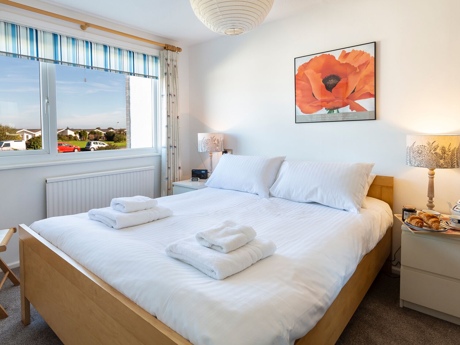 A neatly made bed with white linens and towels, a bedside table with a lamp, a framed artwork on the wall, and a window with curtains.