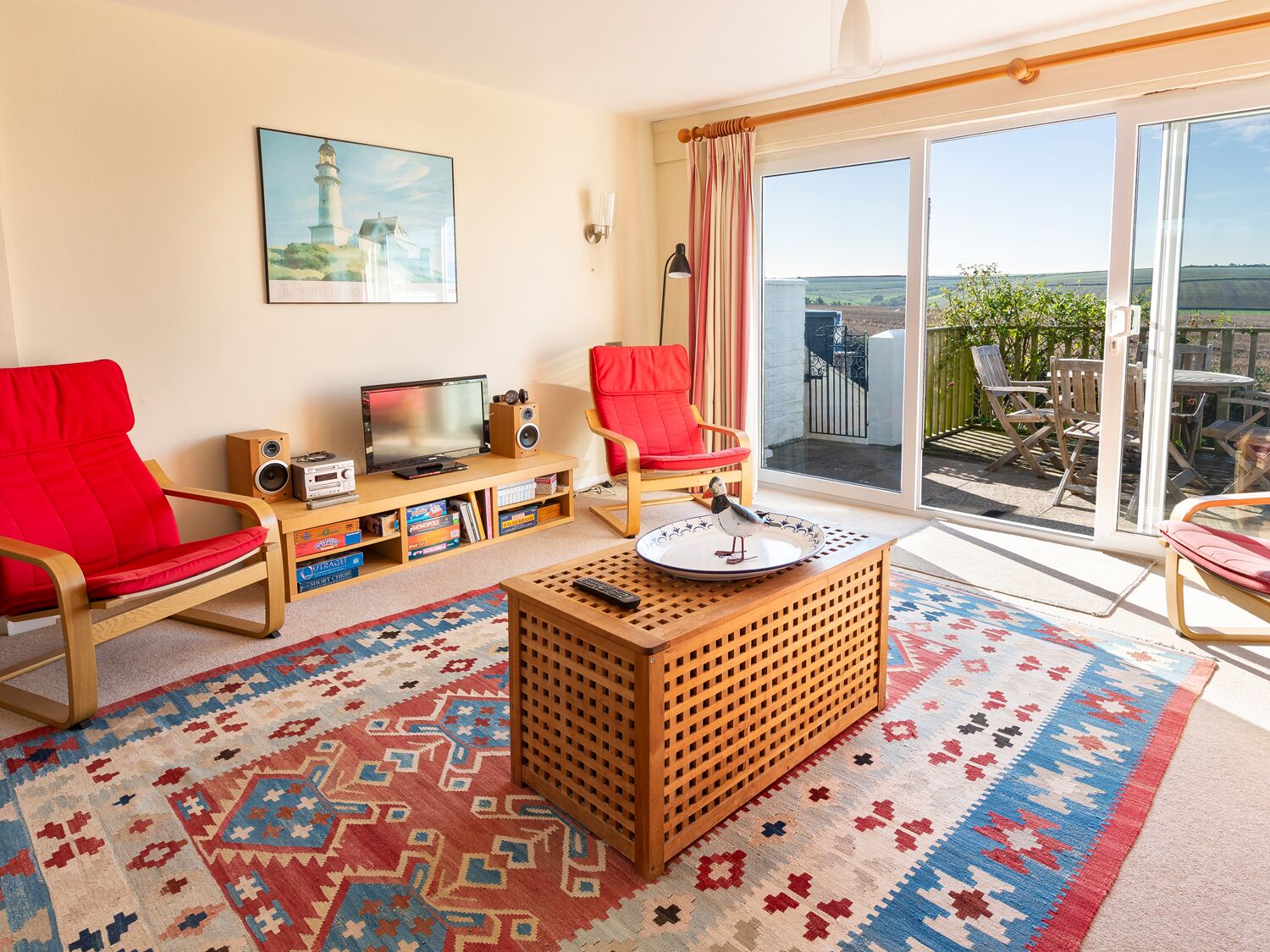 A living room with a TV, speakers, and a red chair. There's a balcony with outdoor furniture and a view of the countryside.