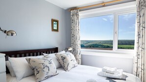 Ein Schlafzimmer mit Bett, Kissen, einer Nachttischlampe, einem Fenster mit Vorhängen und Blick ins Grüne.