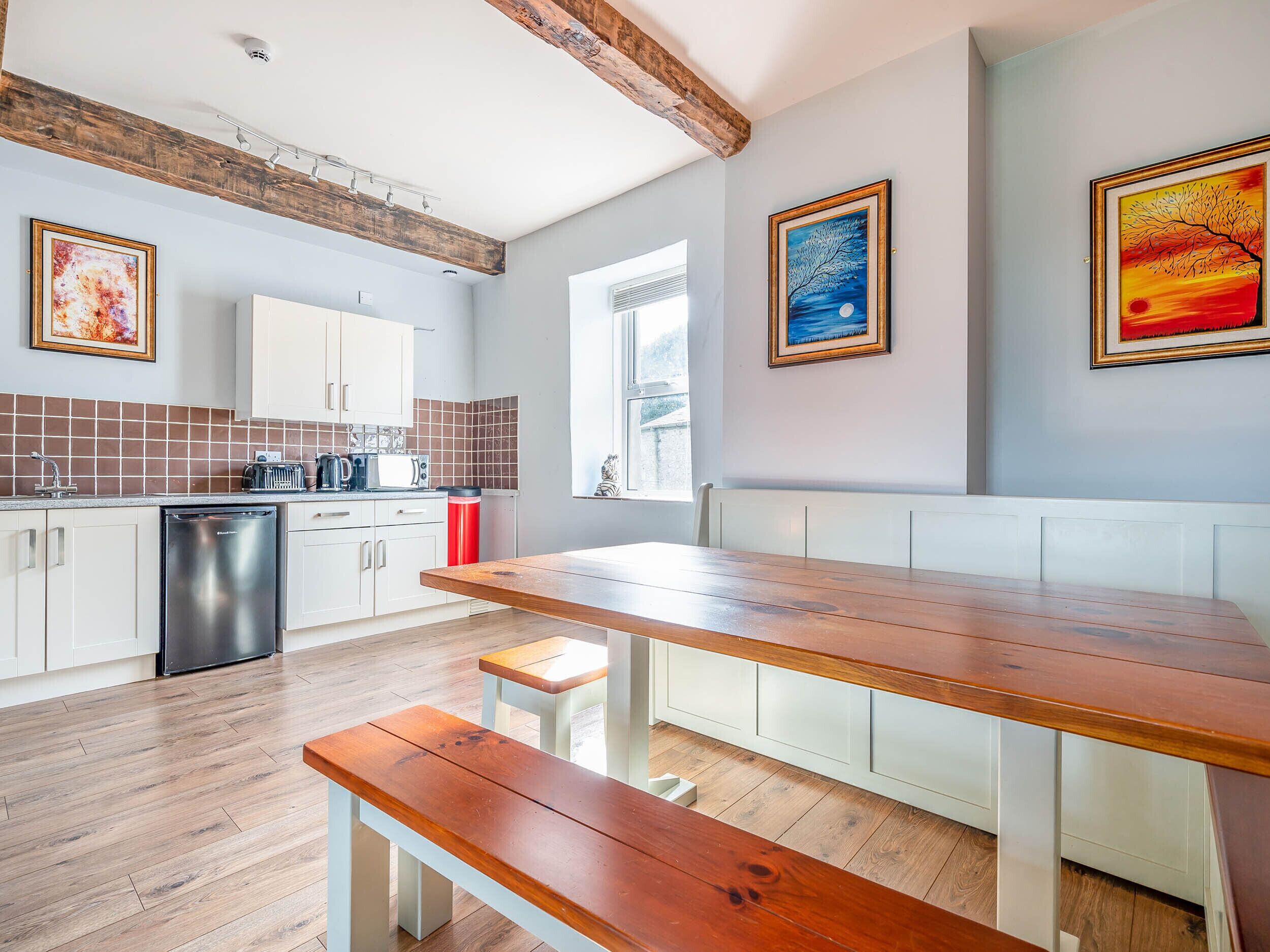 A modern kitchen with white cabinets, a wooden bench, and a dining table. There are three framed artworks on the walls.