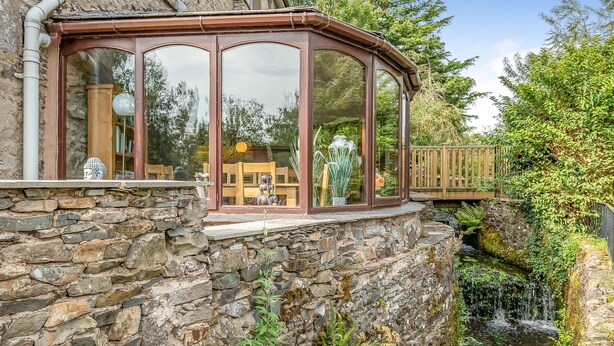 Un bâtiment aux murs de pierre, doté d’une véranda en verre, d’une petite cascade et d’une terrasse en bois.