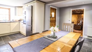 A modern kitchen with a built-in refrigerator, a dining area with a wooden table and chairs, and a stone wall in the background. - Cwm Yr Afon Mawr (Llanbedr)