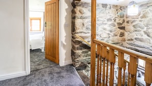 A hallway with a wooden door leading to a bedroom, stone walls, and a wooden staircase with a railing. - Cwm Yr Afon Mawr (Llanbedr)