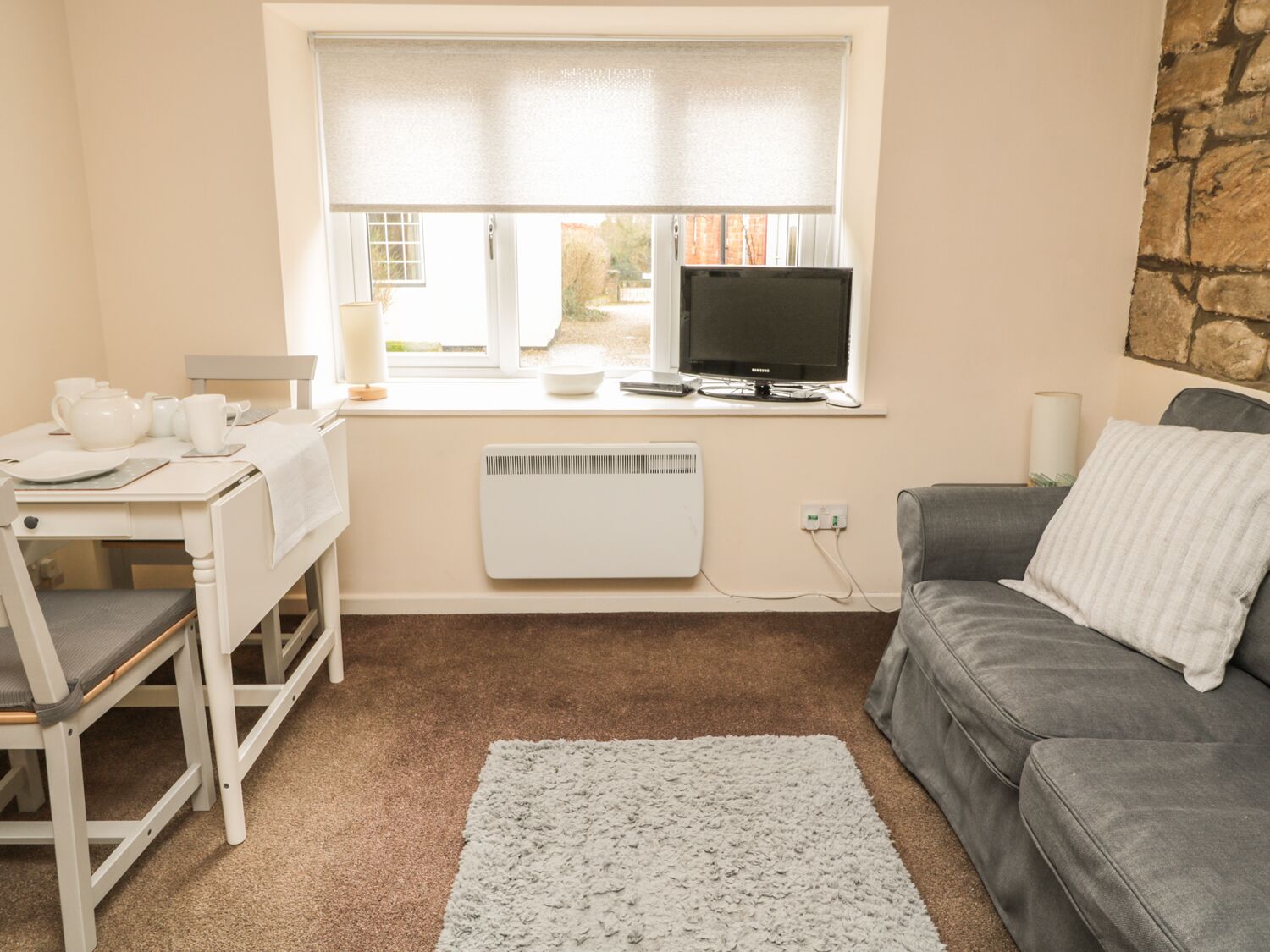 A compact living space with a desk, a grey sofa, a small window with blinds, a radiator, and a television.