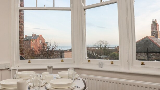A dining area with a table set for four, two windows with views of buildings, and a radiator under the table. - Lovatt House Apartment Tynemouth (North Shields)
