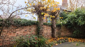 A brick wall with a garden area, including a tree with yellow leaves, a brick building in the background, and a stone pathway. - Lovatt House Apartment Tynemouth (North Shields)
