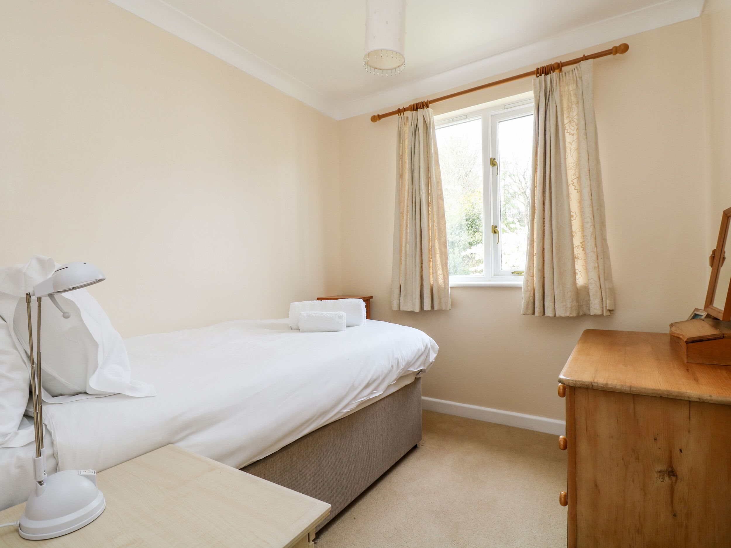 A bedroom with a bed, bedside table, lamp, wooden dresser, and a window with curtains.
