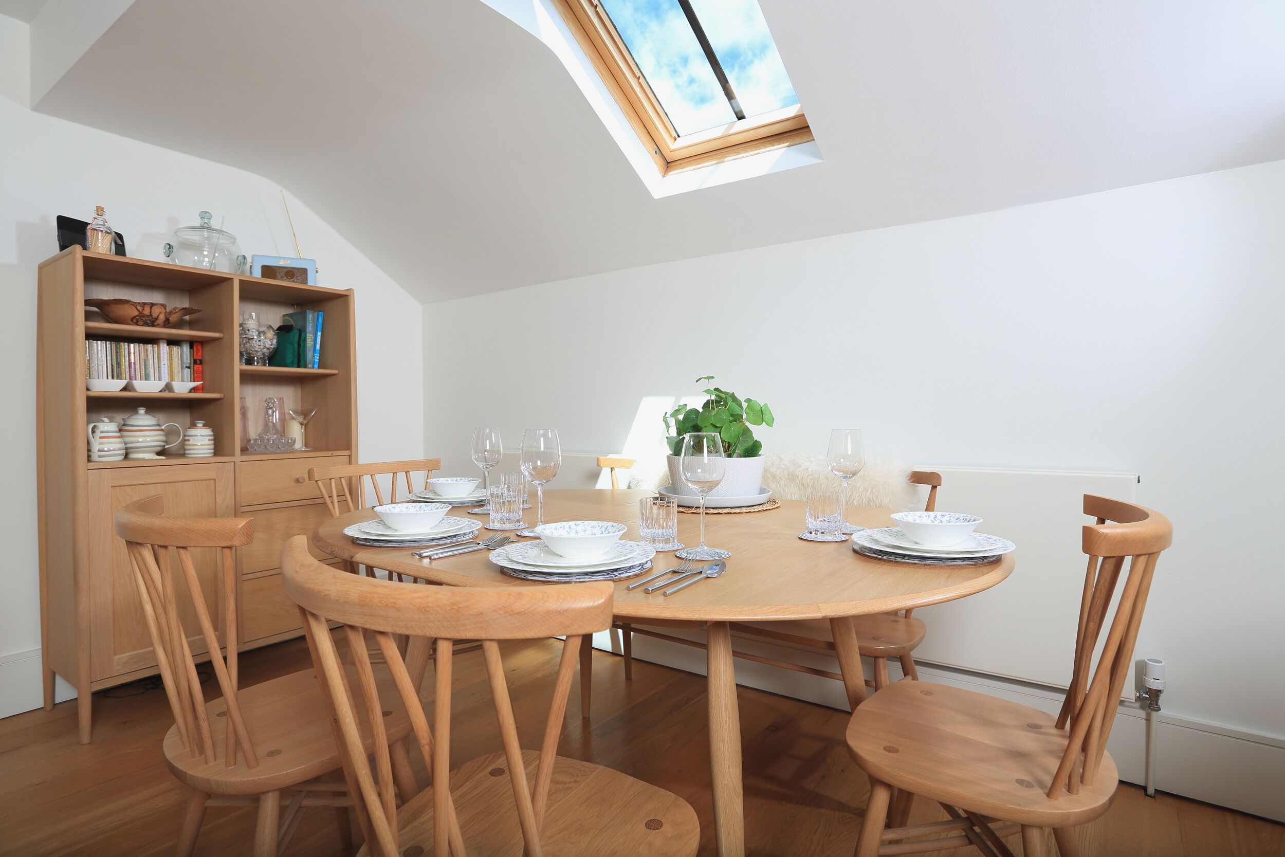 A dining area with a round wooden table set for six, surrounded by wooden chairs. A wooden bookshelf with various items is to the left.