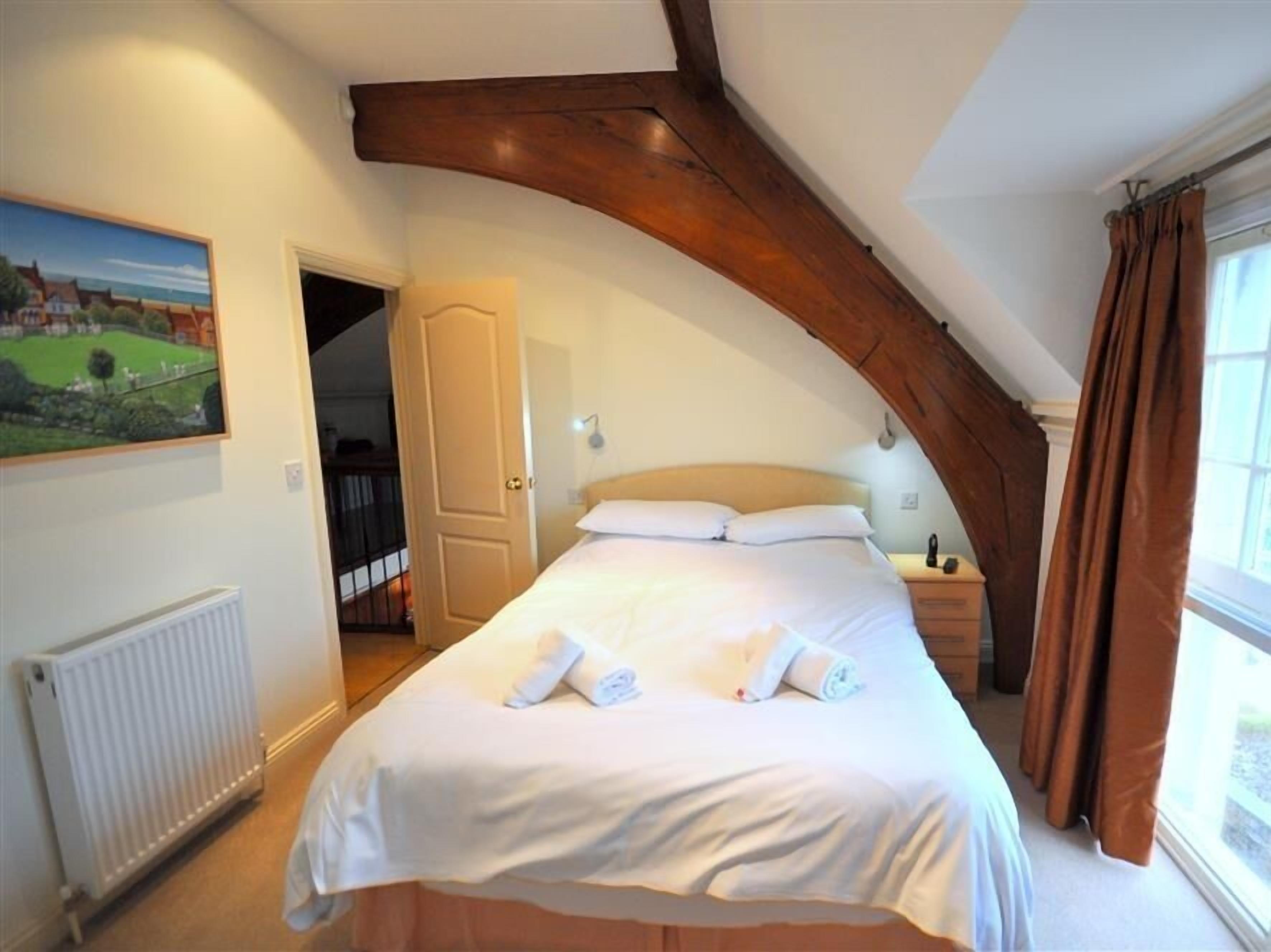 A bedroom with a large bed, a wooden ceiling beam, a radiator, and a window with curtains.