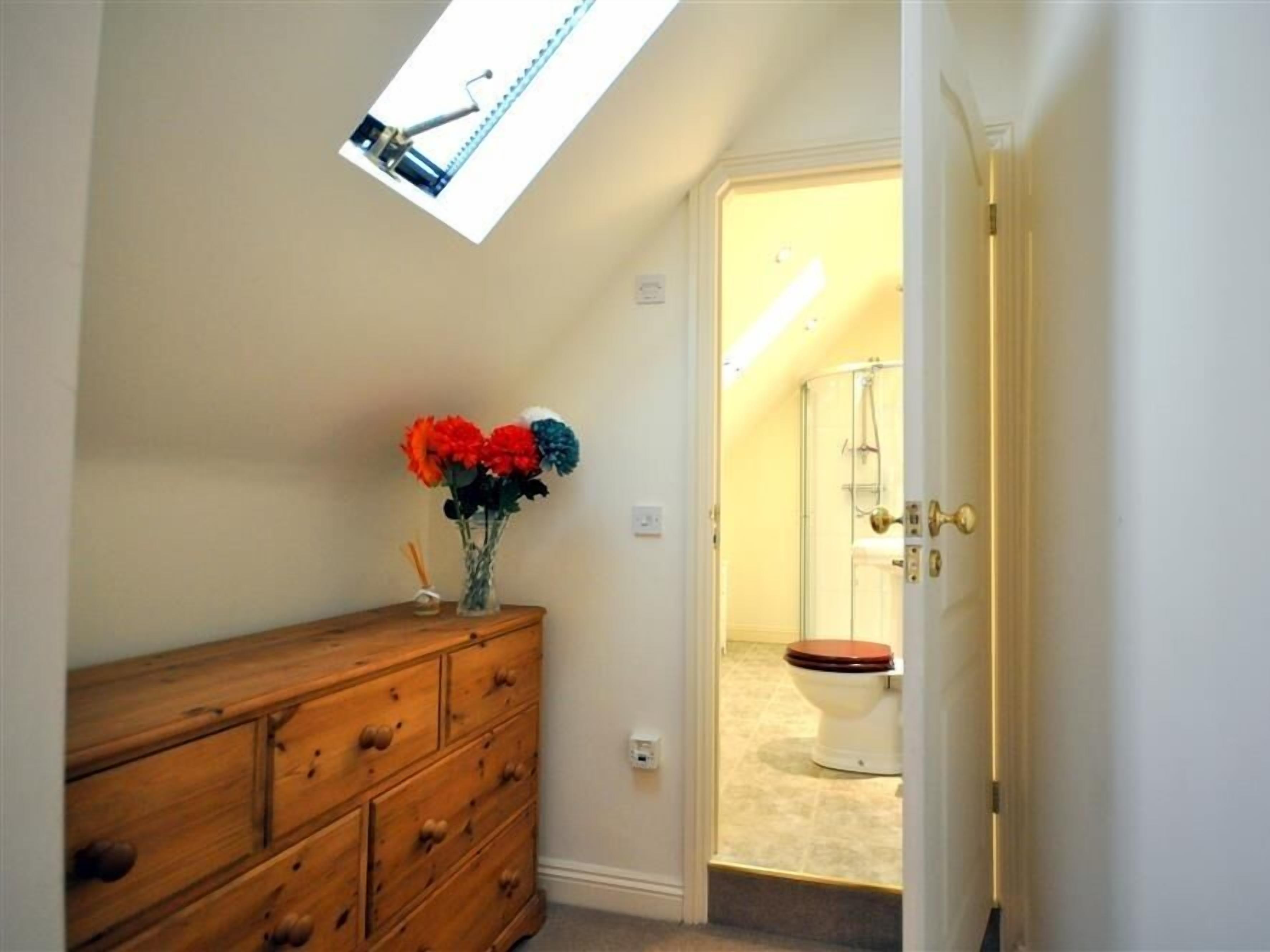 A bathroom with a wooden dresser, a vase with flowers, and a skylight above the toilet.