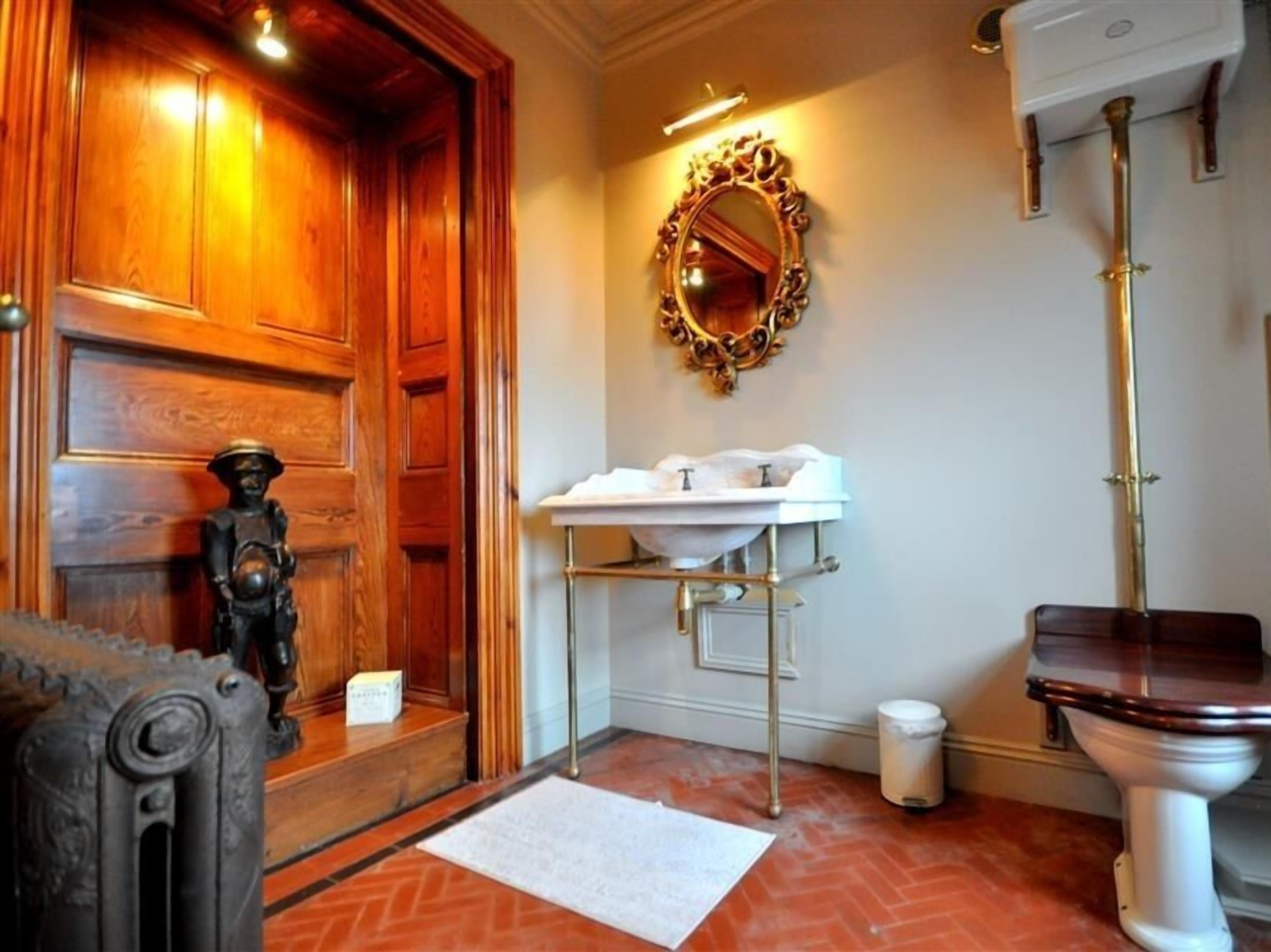 A bathroom with a wooden door, a white sink with a gold faucet, a mirror with an ornate frame, and a toilet.