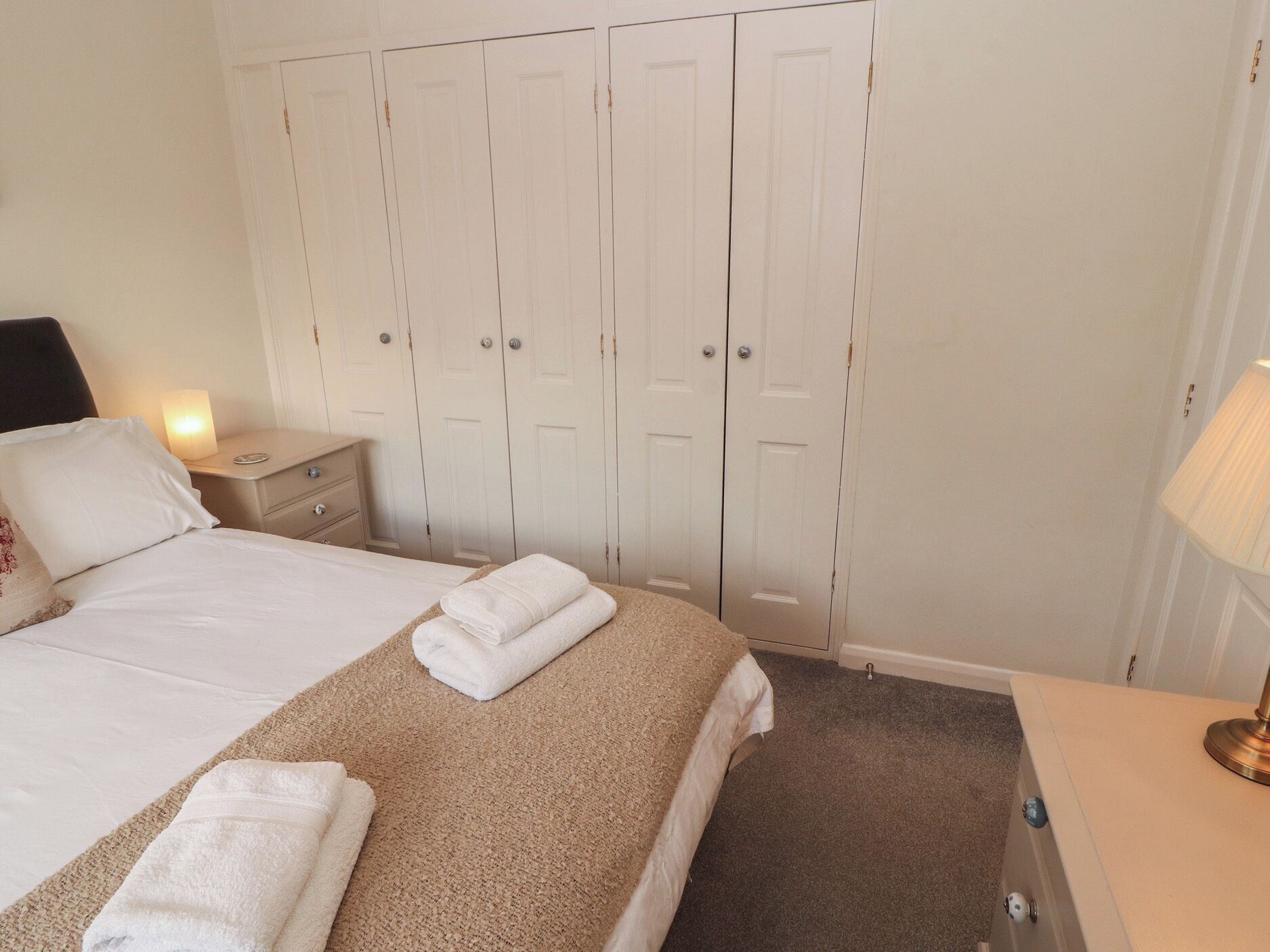 Un lit bien fait, avec du linge de lit blanc et une couverture beige, une lampe de chevet et une table de chevet avec une lampe.