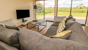 A living room with a large sofa, a coffee table, and a flat-screen TV. There is a large window with a view of a grassy field. - Rose Lodge (York)