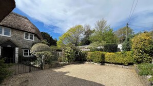 A stone cottage with a thatched roof, a gravel driveway, and a well-maintained garden with various shrubs and trees. - Ivy Cottage (Wareham)