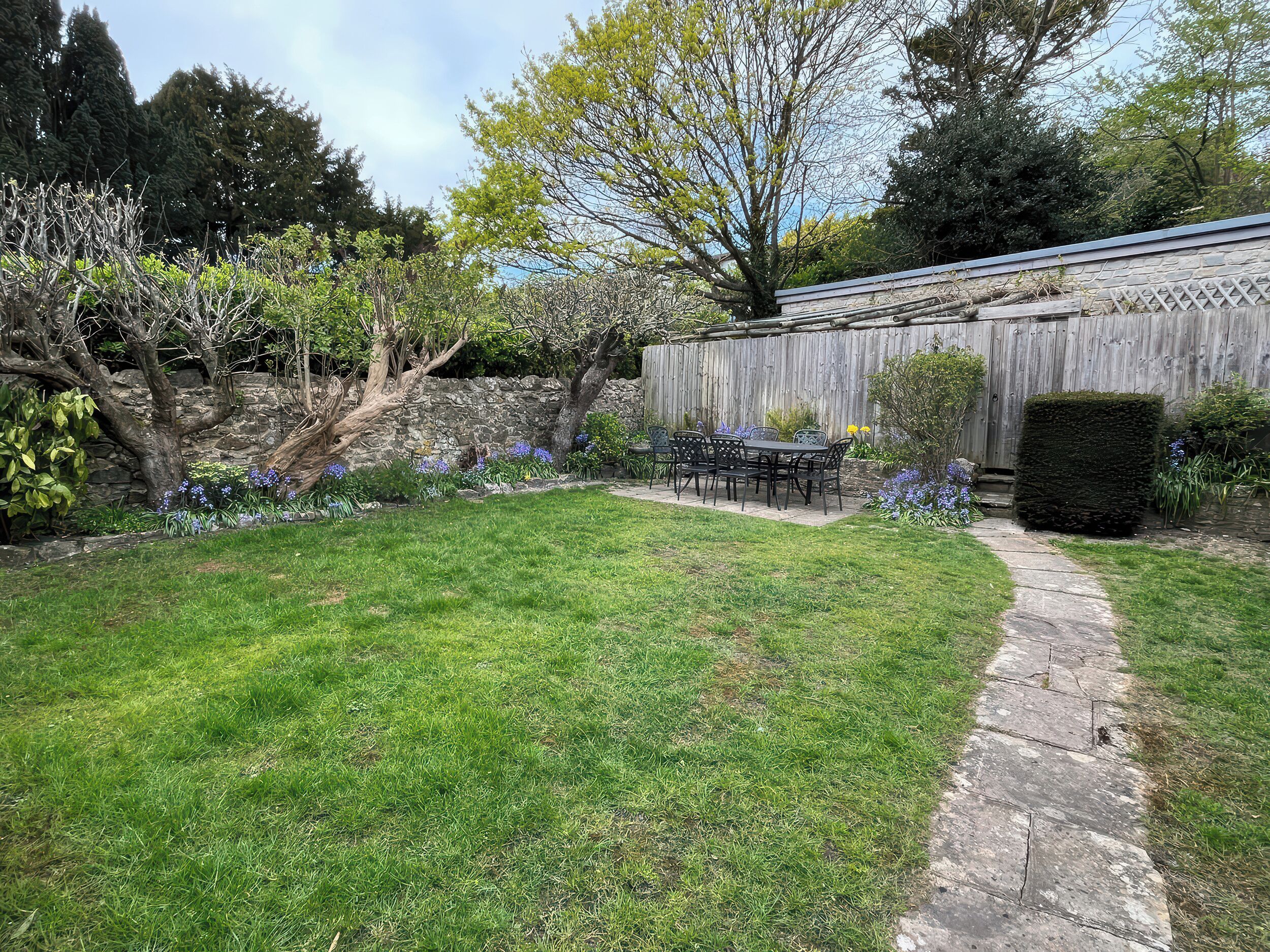 A stone pathway leading to a patio with a table and chairs, surrounded by a grassy yard and flowering plants.