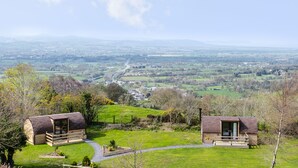 Two rustic cabins with wooden exteriors, set on a grassy hill overlooking a valley with a winding road and distant buildings. - Gelert Pod (Saint Asaph)