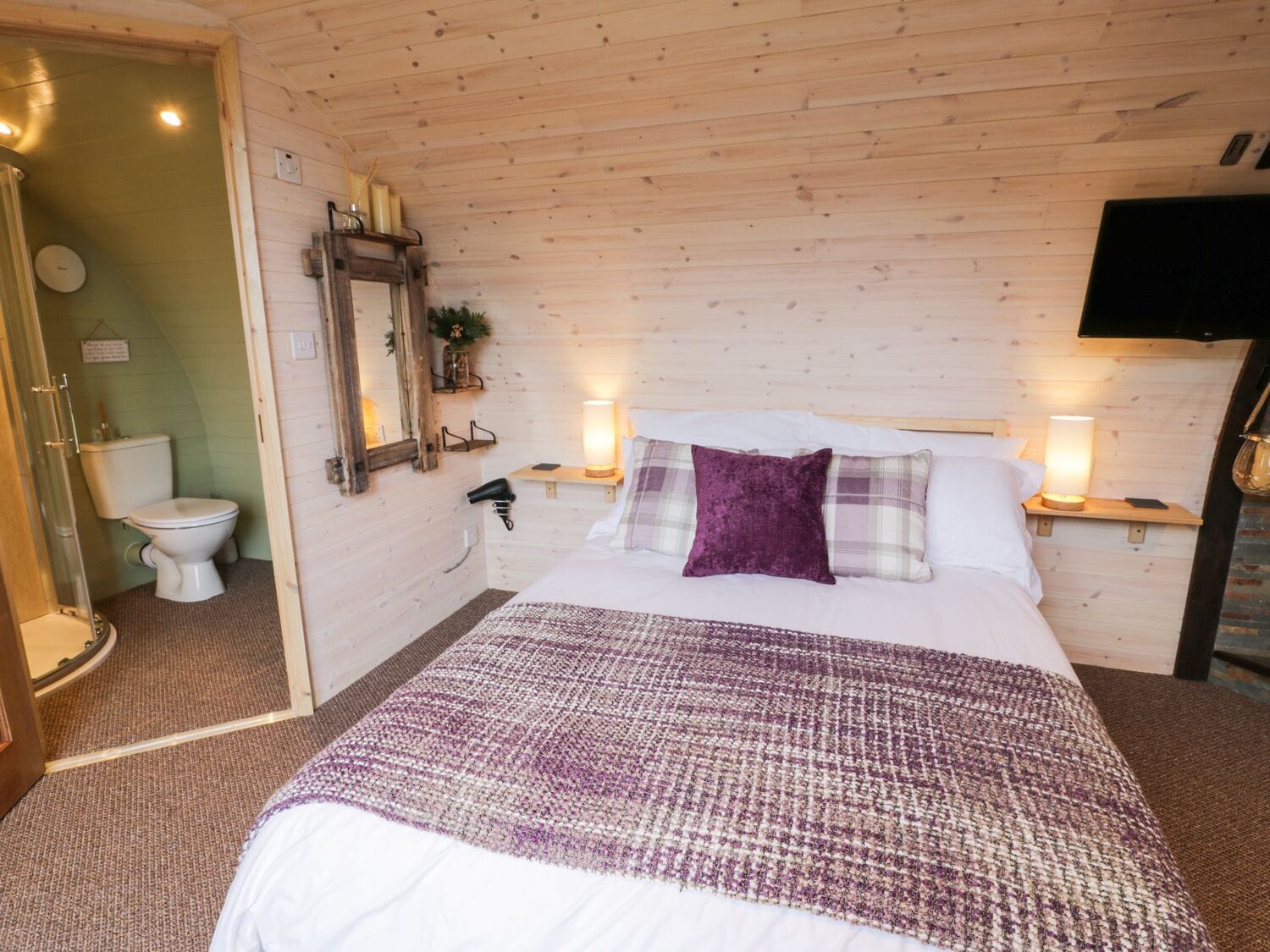 Un dormitorio con techo de madera, una cama con almohada morada, mesitas de noche con lámparas, un televisor montado y un espejo con marco de madera.