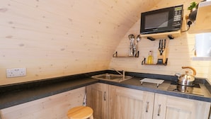 A compact kitchen with wooden cabinets, a black countertop, a microwave, a kettle, and two stools. - Gelert Pod (Saint Asaph)