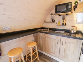 A compact kitchen with wooden cabinets, a black countertop, a microwave, a kettle, and two stools.