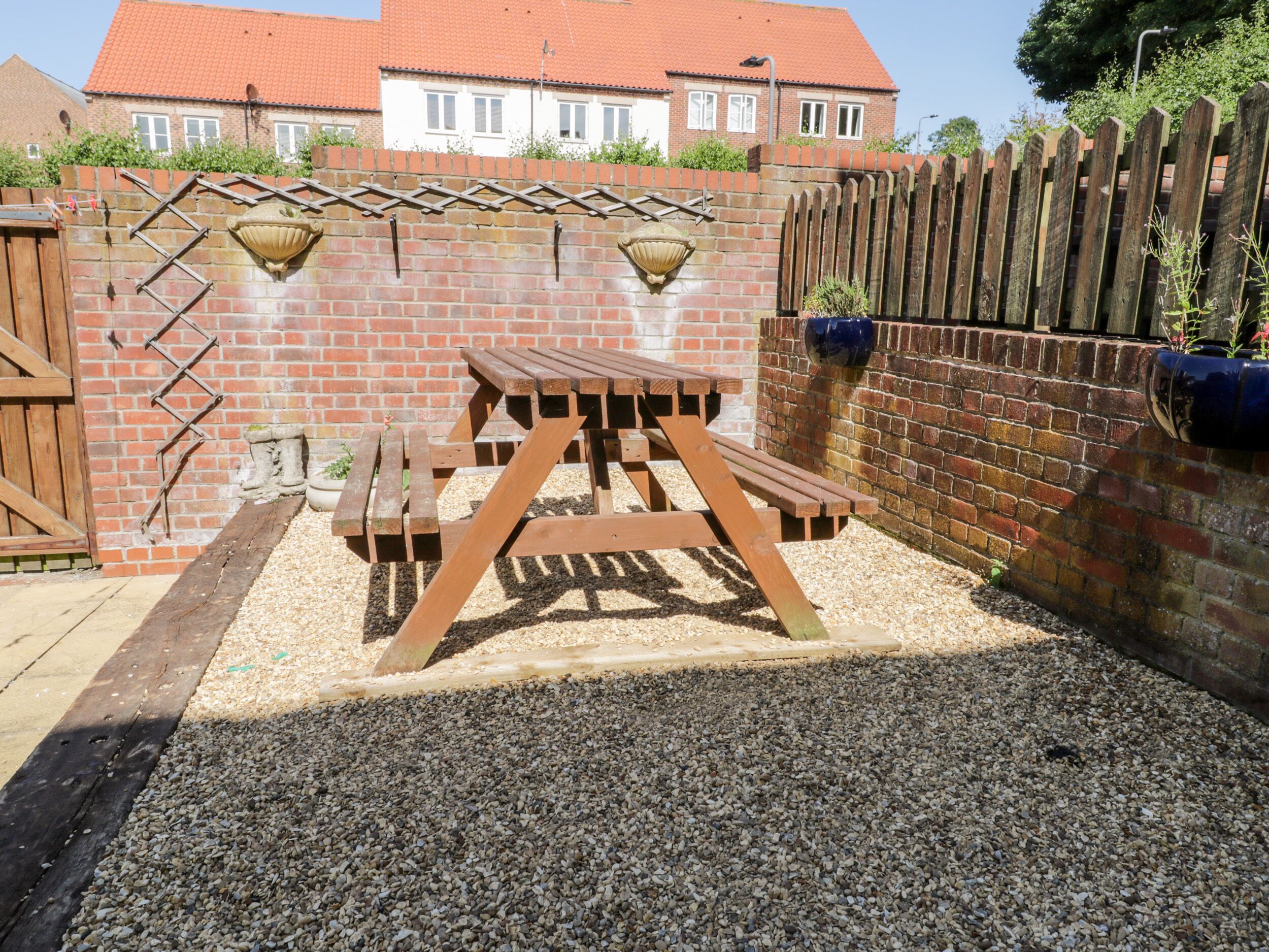 A wooden picnic table with benches in a gravel area, surrounded by a brick wall with decorative elements and a wooden fence.
