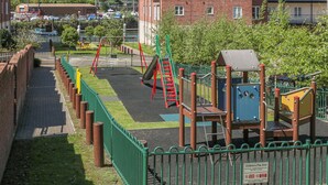 Un parque infantil con columpios y un tobogán, rodeado por una valla verde y un edificio de ladrillo al fondo.