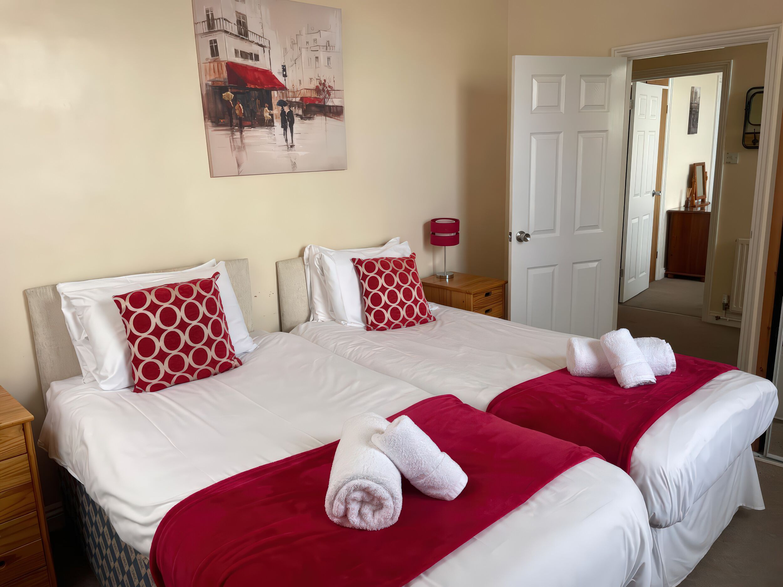 A hotel room with two beds, each with white bedding and red accents, a painting on the wall, and a door leading to another room.